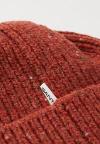 Bonnet en laine tricoté de couleur rouille avec des éclats de vert et de rouge, arborant une étiquette de marque GANT blanche sur le revers.
