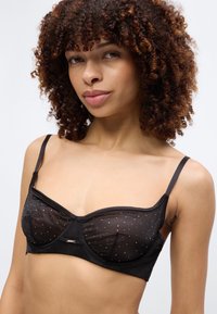 Νεαρή γυναίκα με σγουρά μαλλιά που φοράει μαύρο διάφανο σουτιέν διακοσμημένο με μικρές λαμπερές λεπτομέρειες, ποζάροντας μπροστά σε απλό φόντο.