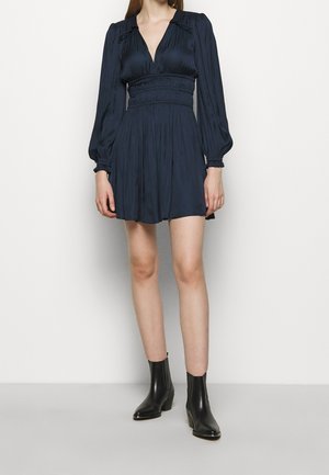 Femme portant une robe courte manches longues bleu marine avec taille et poignets smockés, associée à des bottines noires sur fond blanc uni.