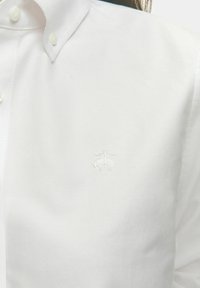 Biała koszula z guzikiem, wykonana z gładkiego materiału, z dyskretnym haftowanym logo na piersi i klasycznym wzornictwem kołnierzyka.