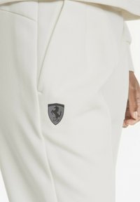 Närbild av vita byxor med en svart prancing horse Ferrari-logotyp nära fickan och en persons hand som vilar vid sidan.