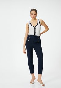 Marineblauwe high-waisted broeken met gouden knopen, gecombineerd met een witte geribbelde tanktop met marineblauwe accent en een diepe V-hals.