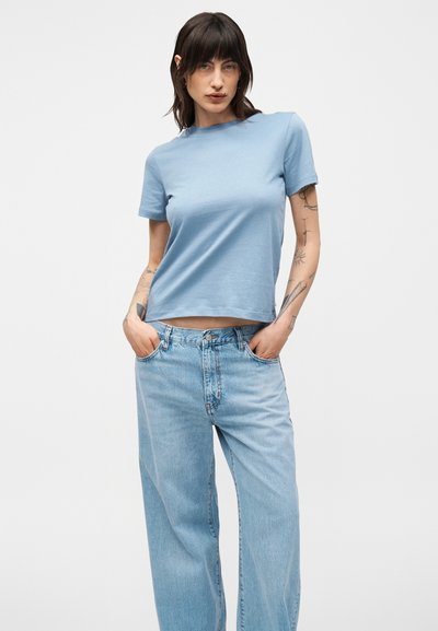 Γυναίκα με σκούρα μαλλιά που φορά ένα ανοιχτό μπλε κοντομάνικο πουκάμισο και άνετο ανοιχτό μπλε τζιν, στέκεται με τα χέρια στις τσέπες σε λευκό φόντο.