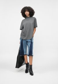 Sivá oversized tričko s krátkymi rukávmi a logom, v kombinácii s nohavicami v patchworkovanom dizajne až po kolená v modrých odtieňoch a čiernymi členkovými topánkami. Čierna plážová taška.