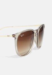 Ray-Ban sunglasses with a transparent frame, brown gradient lenses, and gold metal accents. Features a distinctive logo on the temple.