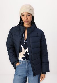 Jachetă de puffer de marină cu textură matlasată, guler înalt și închidere cu fermoar. Asortată cu o cămașă cu model și blugi.