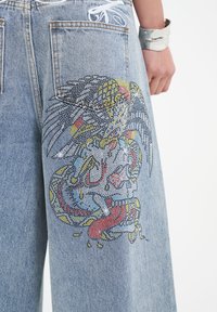 Jasnoniebieskie dżinsy z detalicznym, kolorowym wzorem graficznym na plecach, przedstawiające wzór ptaka i smoka z metalicznymi akcentami.