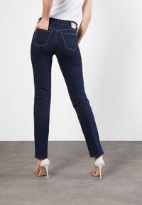 Jeans ajustados de un azul oscuro con dos bolsillos traseros, una cinturilla alta y costuras en contraste, combinados con zapatos de tacón alto blancos.