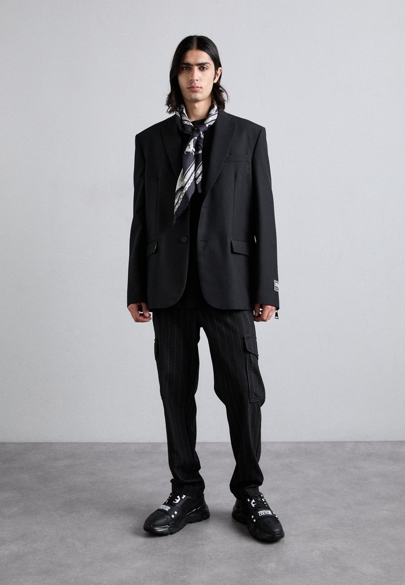 Schwarze, übergroße Blazer mit Knöpfen, kombiniert mit gestreiften schwarzen Cargo-Hosen und schwarzen Sneakers. Ein gemusterter schwarz-weißer Schal akzentuiert das Outfit.