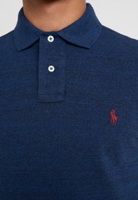 Närbild av en person som bär en blå stickad polo med två knappar och en liten röd broderad polo-spelare-logga.