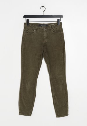 Pantalones ajustados de pana verde oliva colgados en una percha negra contra un fondo blanco.