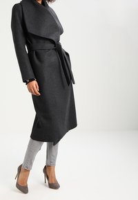 Cappotto in lana grigia con design avvolgente, maniche lunghe, cintura da annodare e taglio fino al ginocchio. Abbinato a jeans grigi e décolleté in tonalità soft.