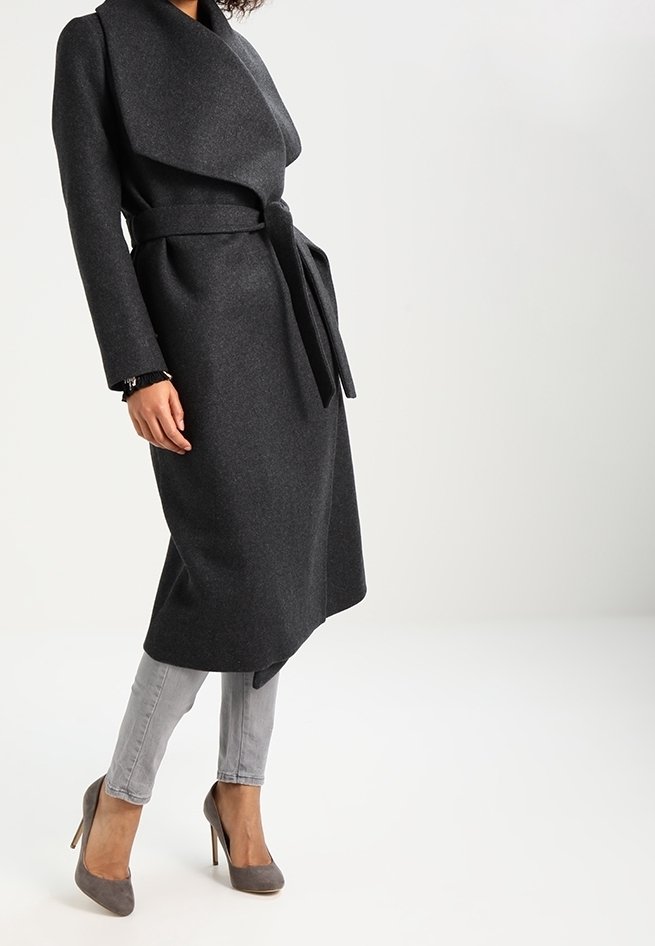 Cappotto in lana grigia con design avvolgente, maniche lunghe, cintura da annodare e taglio fino al ginocchio. Abbinato a jeans grigi e décolleté in tonalità soft.