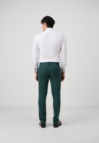 Chemise blanche à manches longues avec des boutons de poignet noirs, associée à un pantalon vert ajusté avec des poches arrière zippées. Texture de tissu lisse.