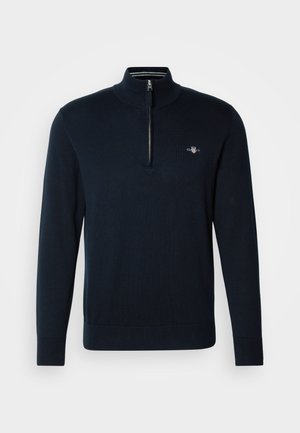 Pull en maille bleu marine avec un col zippé à mi-hauteur, des poignets côtelés et un petit logo brodé sur la poitrine. Texture lisse, coupe décontractée.