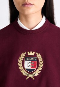 Sudadera color marrón con un emblema bordado que presenta una corona, una guirnalda de laurel y el texto "TOMMY NEW YORK" en el centro. Cuello acanalado clásico.