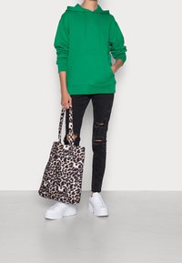 Sudadera verde con un bolsillo canguro, combinada con jeans negros rasgados. La bolsa tote presenta un diseño de estampado de leopardo beige y negro. Zapatillas blancas.