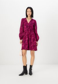 Robe courte à fleurs roses avec une taille cintrée, des manches longues bouffantes et un décolleté en V. Associée à des bottines noires et à un petit sac à main.