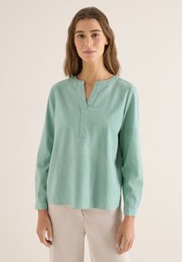 Femme aux cheveux bruns mi-longs portant un chemisier texturé vert clair à manches longues et un pantalon beige sur un fond uni.