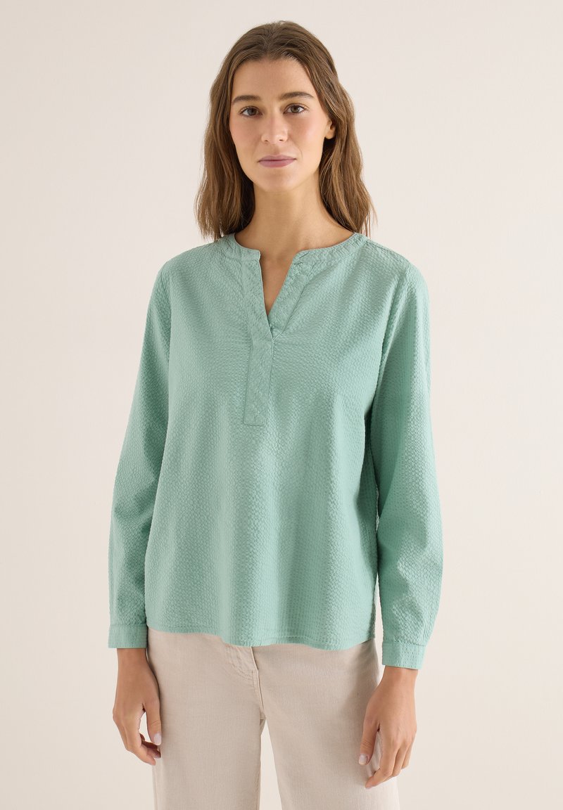 Femme aux cheveux bruns mi-longs portant un chemisier texturé vert clair à manches longues et un pantalon beige sur un fond uni.