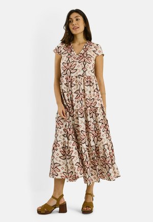 Femme debout portant une robe crème mi-longue avec des motifs floraux rouges et noirs et des sandales à plateforme à talons marron.