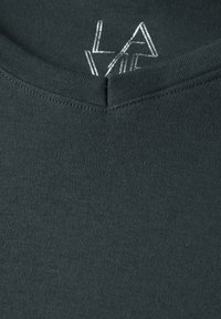 T-shirt vert foncé avec un décolleté en V, fabriqué en coton doux. Présente un petit logo brodé en argent près du col.