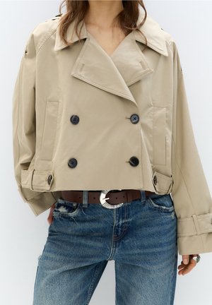 Veste beige courte à double boutonnage avec des boutons noirs portée sur un jean bleu avec une ceinture marron avec une boucle argentée.