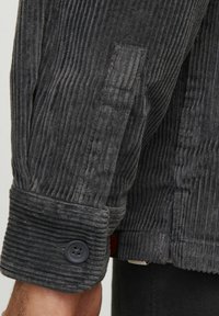 Manschette de veste en velours côtelé gris avec des côtes verticales, fermeture à bouton et finition texturée. Présente un accent contrastant près de la manche.