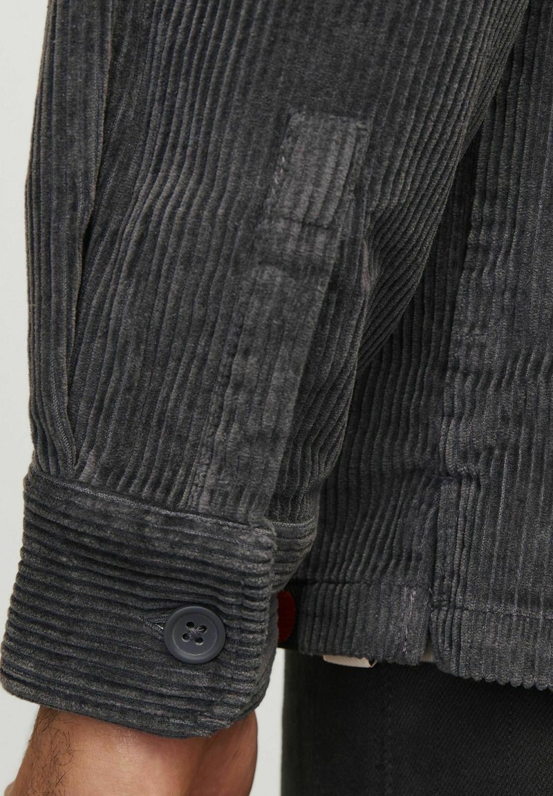 Manschette de veste en velours côtelé gris avec des côtes verticales, fermeture à bouton et finition texturée. Présente un accent contrastant près de la manche.