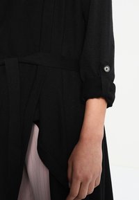 Zwarte gebreide cardigan met een gestructureerde afwerking, een ceintuur om de taille en opgerolde mouwen met een knoopdetail.