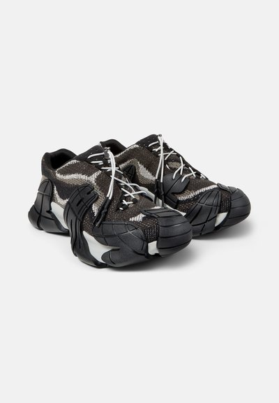 Par sorte og grå futuristiske sneakers med tykke, segmenterede såler og hvide elastiske snørebånd på ensfarvet baggrund.