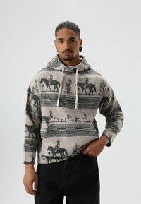 Man draagt een beige en zwarte hoodie met paardenruiter- en cactuspatarnen, hij staat met één hand in de zak tegen een witte achtergrond.