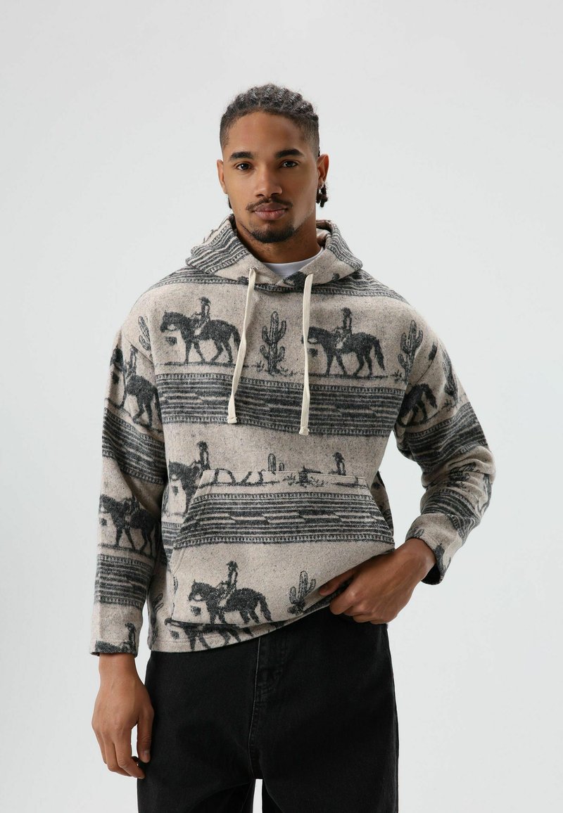 Man draagt een beige en zwarte hoodie met paardenruiter- en cactuspatarnen, hij staat met één hand in de zak tegen een witte achtergrond.