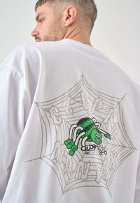 Weißes Baumwoll-T-Shirt mit einem grünen Spinnenmotiv auf einem Netz, umrandet in Grau, auf der Rückseite. Weicher Stoff, lässiges Design.