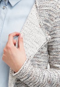 Texturerad vit tröja med grå och bruna inslag, med en lös passform och öppen framsida, vilande mot en ljusblå skjorta.