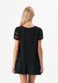 Una donna bionda di spalle che indossa un top nero a maniche corte con maniche di pizzo e una gonna nera plissettata, su uno sfondo bianco semplice.
