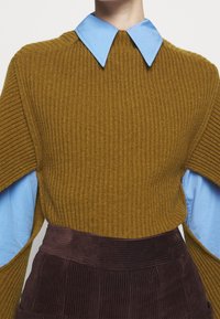 Sininen kaulakotelo ja viiltohihoilla varustettu sinappirikainen neulepaita, yhdistettynä teksturoituun tummanruskeaan corduroy-hameeseen.