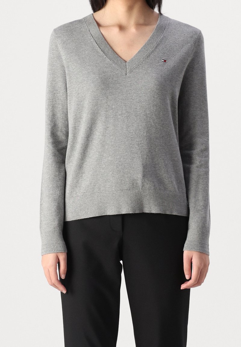 Maglione grigio lavorato a maglia con scollo a V, maniche lunghe e polsini a costine; presenta un piccolo logo sul petto sinistro, abbinato a pantaloni neri.