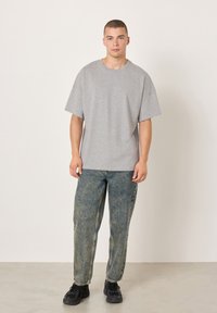 Šedé tričko s krátkým rukávem a volným střihem, doplněné o vybledlé modré džíny s rovnými nohavicemi a černé tenisky na neutrálním pozadí.