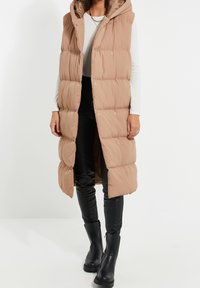 Doudoune sans manches beige clair avec capuche, dotée d'un design matelassé, de boutons-pression et d'ouvertures latérales, associée à un pantalon en cuir noir et des bottes.