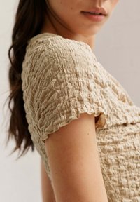 Haut texturé beige clair avec un design de tissu froncé. Manches courtes avec des bords volants, mettant en évidence une apparence douce et légèrement froissée.