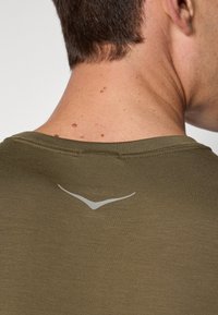 Camiseta verde-oliva com decote redondo, apresentando um pequeno logotipo refletor nas costas, textura suave e mangas curtas.