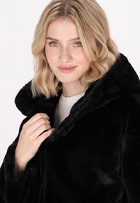 Cappotto in finta pelliccia nera con ampio colletto, texture morbida e vestibilità comoda. La modella tiene il colletto e indossa una maglietta di colore chiaro sotto.