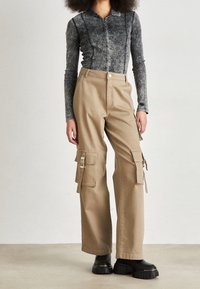 Dlouhý rukáv, šedá džínová košile s knoflíkovým zapínáním, spojena s béžovými cargo kalhotami s více kapsami a nastavovacími bočními popruhy. Černé boty s robustní podrážkou.