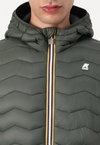 Green padded jacket with a chevron pattern, featuring a zipper with multicolored stripes and a logo on the chest. Hooded design.