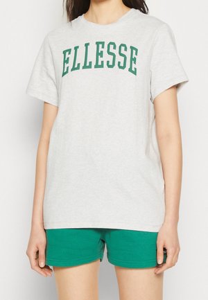 Kobieta w jasnoszarej koszulce z zielonym logo "ELLESSE" i zielonych szortach, stojąca z rękami swobodnie opuszczonymi wzdłuż ciała.
