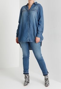 Chemise en denim à manches longues, fermeture à boutons et col pointu. Associée à un jean slim et des bottines à imprimé serpent. Tons bleus prédominants.