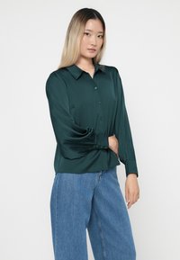 Smaragdgrønn bluse laget av glatt stoff, med knappestolpe foran, spiss krage og lange, løse ermer med knappe- mansjetter.