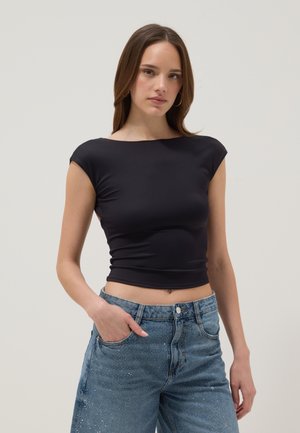 Mujer con una camiseta negra ajustada de manga corta tipo crop top y vaqueros azules de cintura alta, con una mano en el bolsillo, sobre un fondo liso.