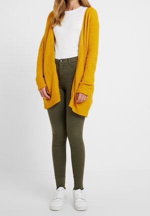 Mujer con cárdigan abierto de color mostaza, camiseta blanca, pantalones pitillo verde oliva y zapatillas blancas, de pie frente a un fondo liso.